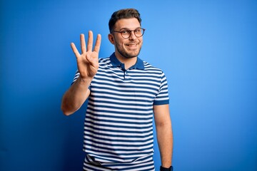 Naklejka premium Young man with blue eyes wearing glasses and casual striped t-shirt over blue background showing and pointing up with fingers number four while smiling confident and happy.