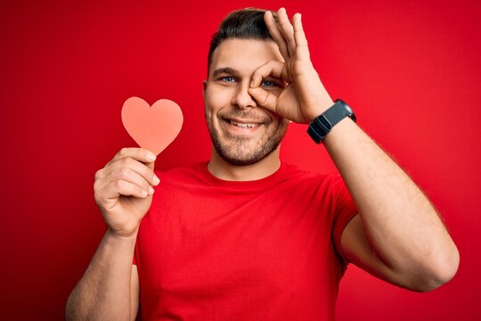 Young romantic man with blue eyes holding red heart paper shaped over red background with happy face smiling doing ok sign with hand on eye looking through fingers