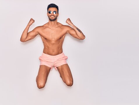 Young handsome hispanic man on vacation wearing swimwear shirtless smiling happy. Jumping with smile on face celebrating with fists up over isolated white background