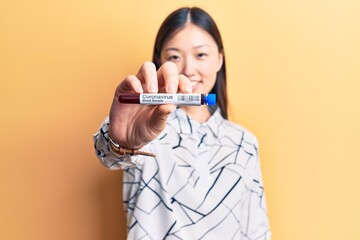 Young beautiful chinese woman holding test tube with coronavirus blood sample looking positive and happy standing and smiling with a confident smile showing teeth