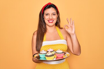 Young hispanic pin up woman wearing fashion sexy 50s style holding sweet cupcakes doing ok sign with fingers, excellent symbol