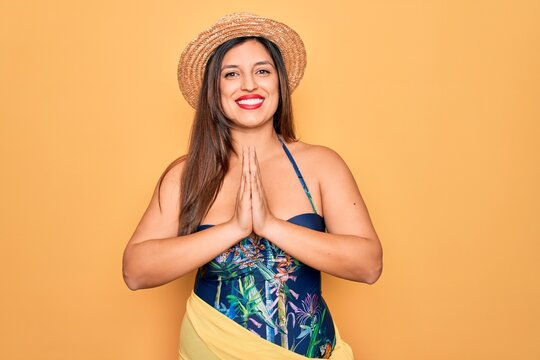Young hispanic woman wearing summer hat and swimsuit over yellow background praying with hands together asking for forgiveness smiling confident.