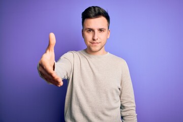 Naklejka premium Young handsome caucasian man wearing casual sweater over purple isolated background smiling friendly offering handshake as greeting and welcoming. Successful business.