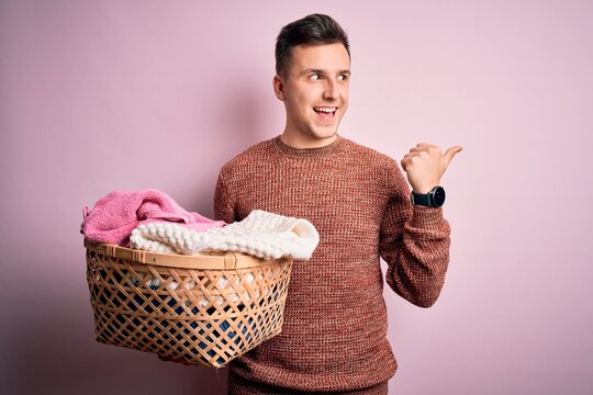 Young handsome caucasian man doing housework chores holding laundry wicker basket pointing and showing with thumb up to the side with happy face smiling