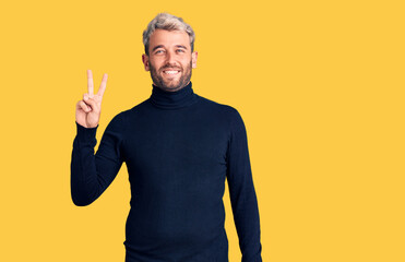 Young handsome blond man wearing casual turtleneck sweater smiling with happy face winking at the camera doing victory sign. number two.