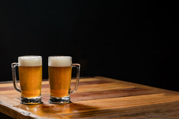 two cold mugs with beer, with overflowing foam, on wooden table and dark background, space for writing