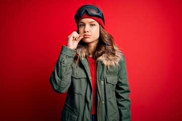 Young blonde girl wearing ski glasses and winter coat for ski weather over red background mouth and...