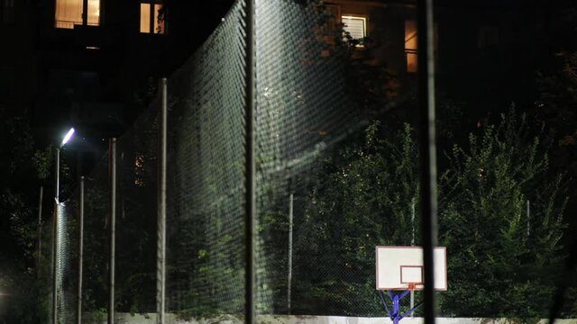 Standing at fence of large inner-city basketball court at night. Big modern sports playground lighted by street lights in empty neighbourhood. Sport and active lifestyle concept.