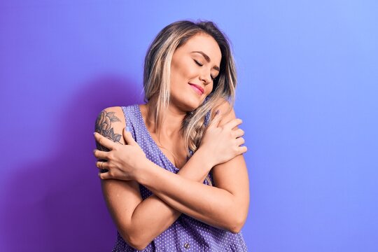 Young beautiful blonde woman wearing casual t-shirt standing over isolated purple background hugging oneself happy and positive, smiling confident. Self love and self care
