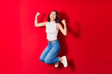 Young beautiful chinese woman wearing casual clothes smiling happy. Jumping with smile on face doing winner gesture with fists up over isolated red background