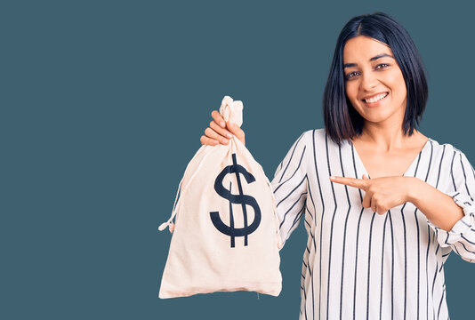 Young beautiful latin woman holding money bag with dollar symbol smiling happy pointing with hand and finger