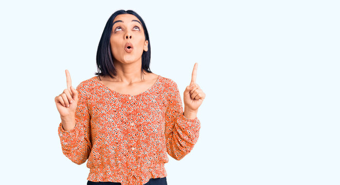 Young beautiful latin woman wearing casual clothes amazed and surprised looking up and pointing with fingers and raised arms.