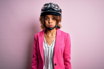 African american motorcyclist woman with curly hair wearing moto helmet over pink background puffing cheeks with funny face. Mouth inflated with air, crazy expression.