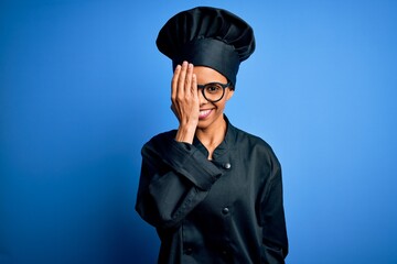 Young african american chef woman wearing cooker uniform and hat over blue background covering one eye with hand, confident smile on face and surprise emotion.