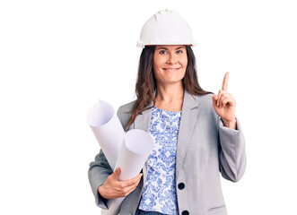 Young beautiful brunette woman wearing architect hardhat holding blueprint surprised with an idea or question pointing finger with happy face, number one