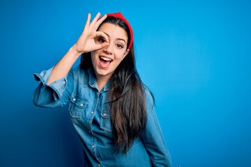 Young brunette woman wearing casual denim shirt over blue isolated background doing ok gesture with hand smiling, eye looking through fingers with happy face.