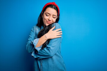 Young brunette woman wearing casual denim shirt over blue isolated background Hugging oneself happy and positive, smiling confident. Self love and self care