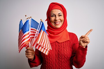 Middle age patriotic woman wearing traditional muslim hijab holding united states flags very happy pointing with hand and finger to the side