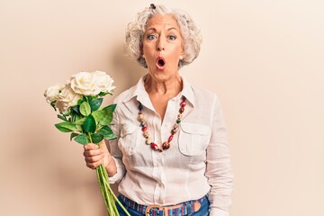 Senior grey-haired woman holding flowers scared and amazed with open mouth for surprise, disbelief...