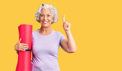 Senior grey-haired woman holding yoga mat surprised with an idea or question pointing finger with...