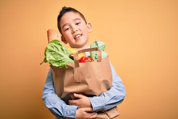 Adorable toddler holding paper bag with food standing over isolated yellow background