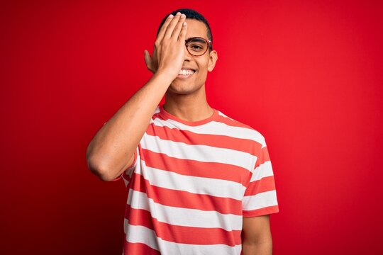 Young handsome african american man wearing casual striped t-shirt and glasses covering one eye with hand, confident smile on face and surprise emotion.