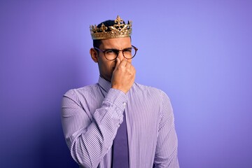 Young handsome african american man wearing golden crown of king over purple background smelling something stinky and disgusting, intolerable smell, holding breath with fingers on nose. Bad smell