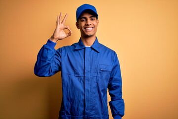 Young african american mechanic man wearing blue uniform and cap over yellow background smiling...