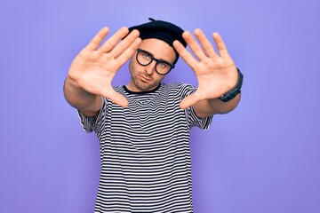 Handsome man with blue eyes wearing striped t-shirt and french beret over purple background doing frame using hands palms and fingers, camera perspective