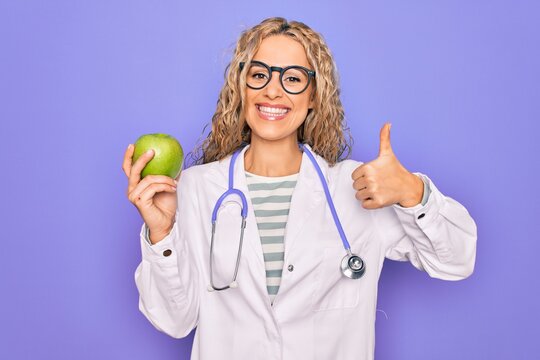Young beautiful blonde doctor woman wearing stethoscope holding green apple fruit Smiling happy and positive, thumb up doing excellent and approval sign