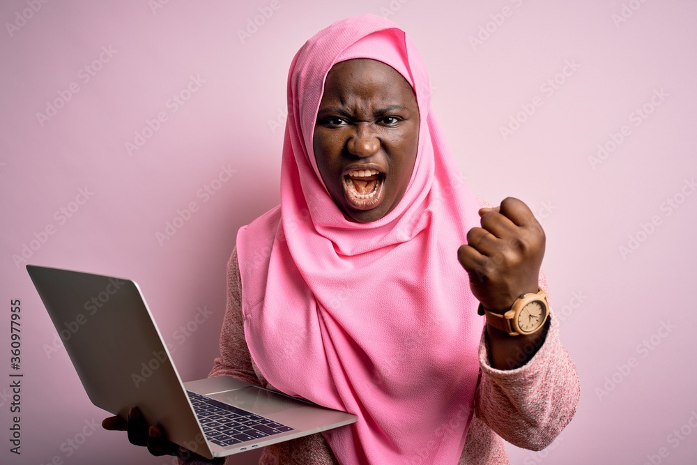 Sticker African american plus size woman wearing muslim hijab using laptop over pink background angry and mad raising fist frustrated and furious while shouting with anger. Rage and aggressive concept.