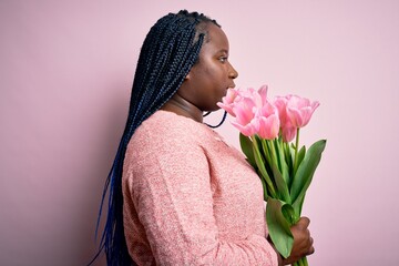 Young african american plus size woman with braids holding bouquet of pink tulips flower looking to side, relax profile pose with natural face and confident smile.