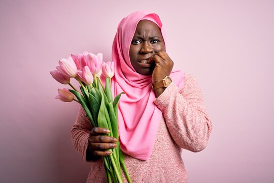 Young african american plus size woman wearing muslim hijab holding bouquet of pink tulips looking stressed and nervous with hands on mouth biting nails. Anxiety problem.
