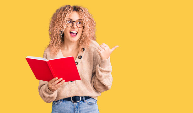 Young blonde woman with curly hair wearing glasses and reading book pointing thumb up to the side smiling happy with open mouth