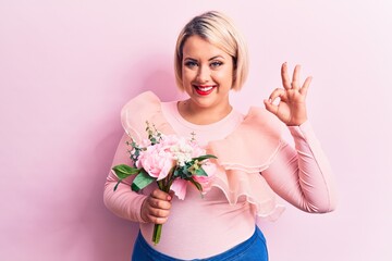 Beautiful blonde plus size woman holding bouquet of pink flowers over isolated background doing ok sign with fingers, smiling friendly gesturing excellent symbol