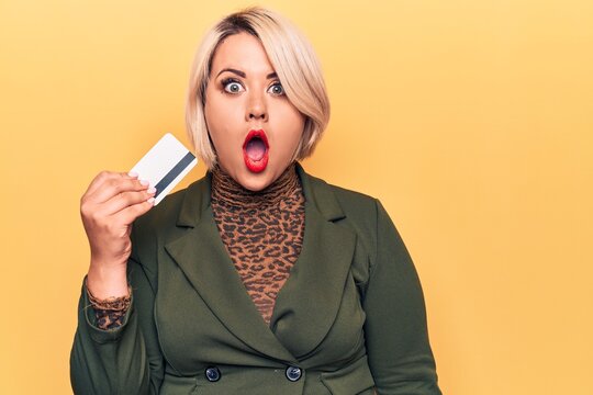 Young beautiful blonde plus size woman holding credit card over isolated yellow background scared and amazed with open mouth for surprise, disbelief face