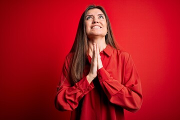 Young beautiful woman with blue eyes wearing casual shirt standing over red background begging and praying with hands together with hope expression on face very emotional and worried. Begging.