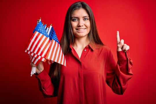 Young beautiful patriotic woman with blue eyes holding united states flags over red background surprised with an idea or question pointing finger with happy face, number one