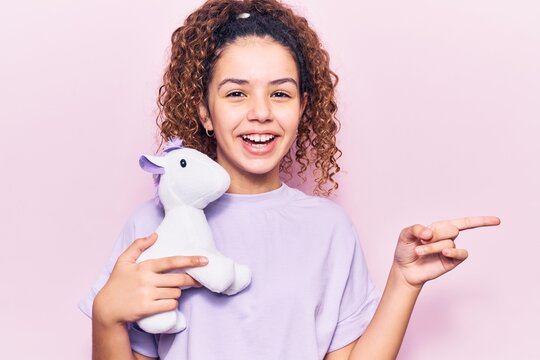 Beautiful kid girl with curly hair holding animal doll toy smiling happy pointing with hand and finger to the side