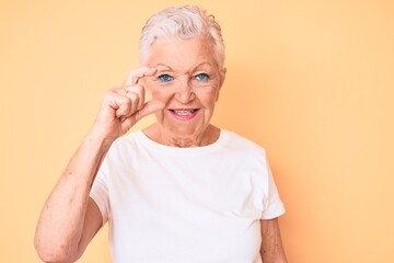 Senior beautiful woman with blue eyes and grey hair wearing classic white tshirt over yellow background smiling and confident gesturing with hand doing small size sign with fingers