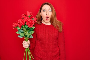 Young beautiful redhead woman holding bouquet of red roses flowers over isolated background scared in shock with a surprise face, afraid and excited with fear expression