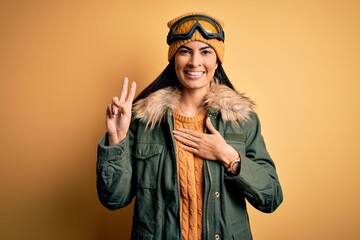 Young beautiful hispanic woman wearing ski glasses and coat for winter weather smiling swearing with hand on chest and fingers up, making a loyalty promise oath
