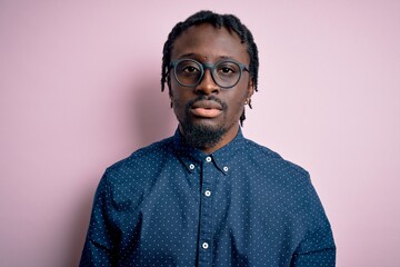 Young handsome african american man wearing casual shirt and glasses over pink background with serious expression on face. Simple and natural looking at the camera.