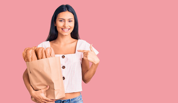 Young beautiful latin girl holding paper bag with bread pointing finger to one self smiling happy and proud