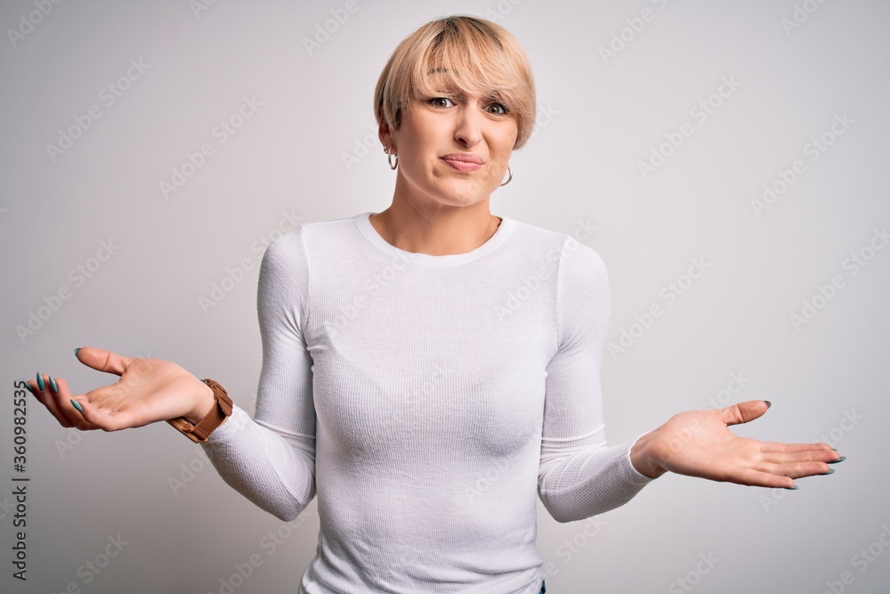 Sticker Young beautiful blonde woman with modern short hair hairstyle standing over isolated background clueless and confused expression with arms and hands raised. Doubt concept.