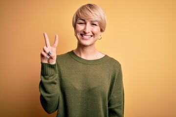 Young blonde woman with modern short hair wearing casual sweater over yellow background showing and pointing up with fingers number two while smiling confident and happy.