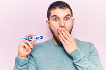 Young handsome man holding thermometer covering mouth with hand, shocked and afraid for mistake....