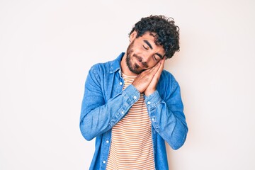 Handsome young man with curly hair and bear wearing casual denim jacket sleeping tired dreaming and posing with hands together while smiling with closed eyes.