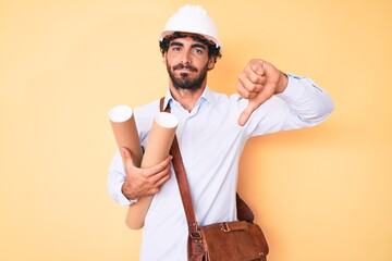 Handsome young man with curly hair and bear holding paper blueprints wearing hardhat with angry...