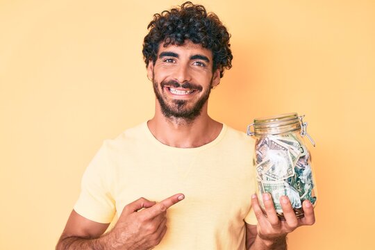 Handsome young man with curly hair and bear holding jar with savings smiling happy pointing with hand and finger
